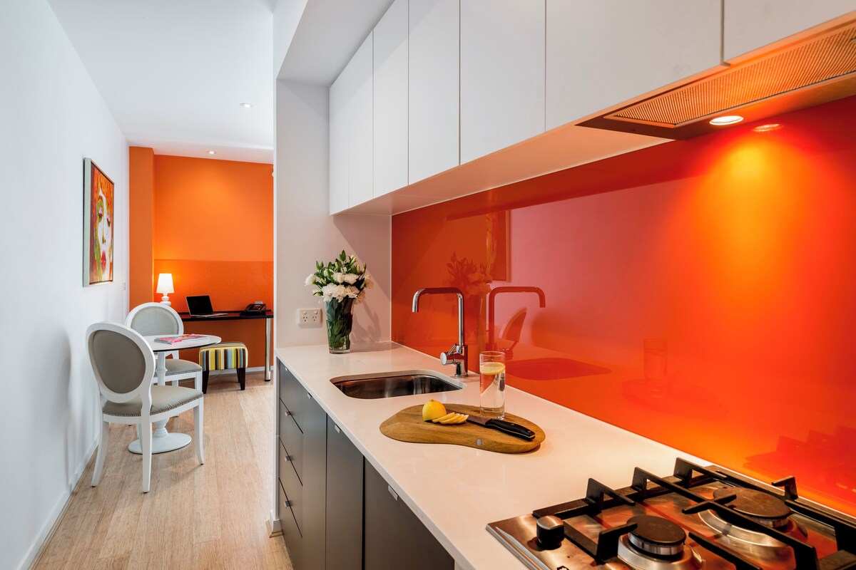 A modern kitchen with a white countertop, a gas stove, and a sink. There is a dining area with a white chair and a small table.