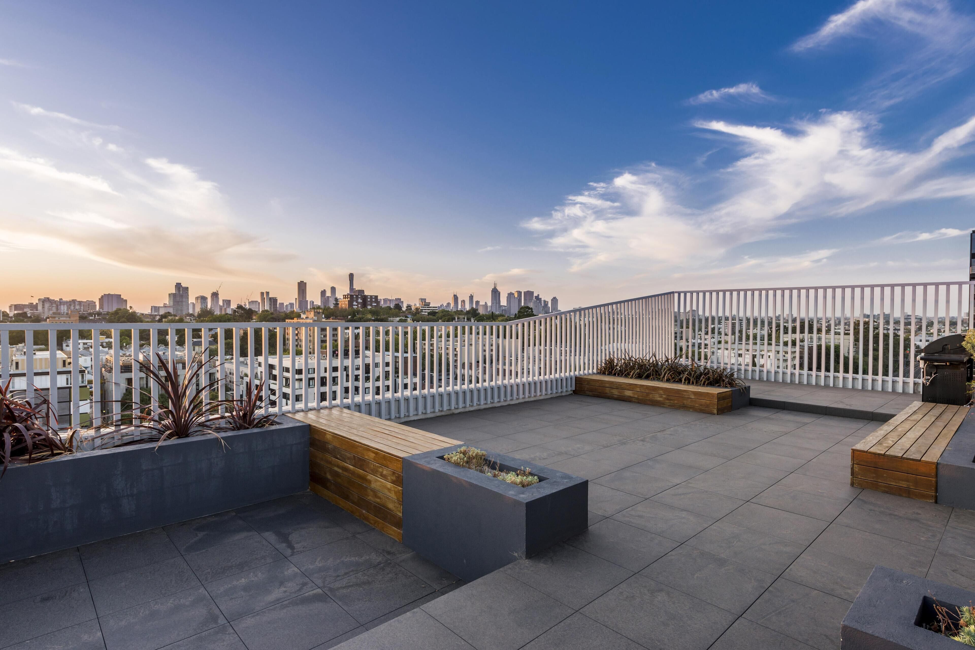 A rooftop terrace with a city skyline view, a white fence, and planters with plants.