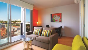 Una sala de estar moderna con un sofá, un sillón amarillo, una mesa de centro de vidrio, una mesa de comedor con sillas y un ventanal con vistas a la ciudad.