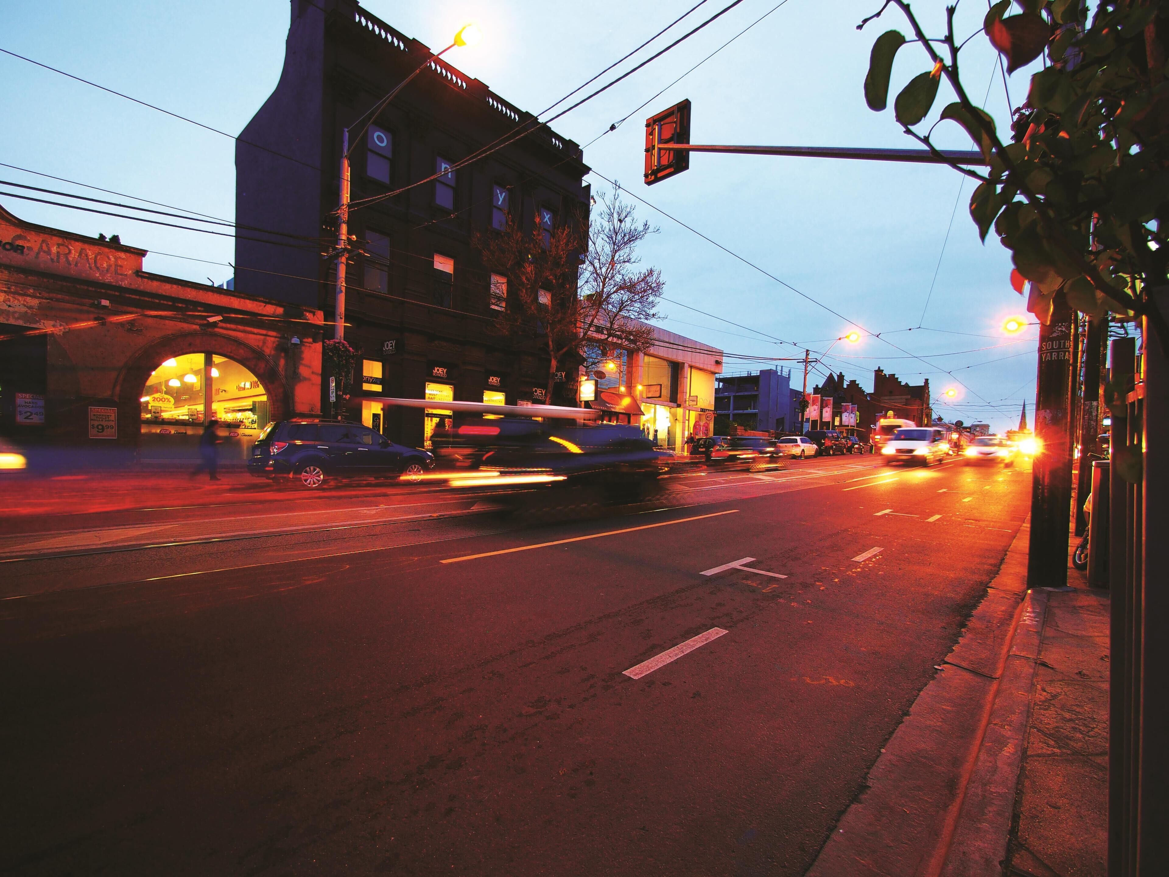 A street view at dusk with buildings, a pedestrian crossing, and traffic lights.