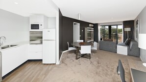 Een moderne keuken met witte kastjes, een magnetron en een koelkast. Er is een zithoek met een tafel en stoelen, en een woongedeelte met een bank en een televisie.