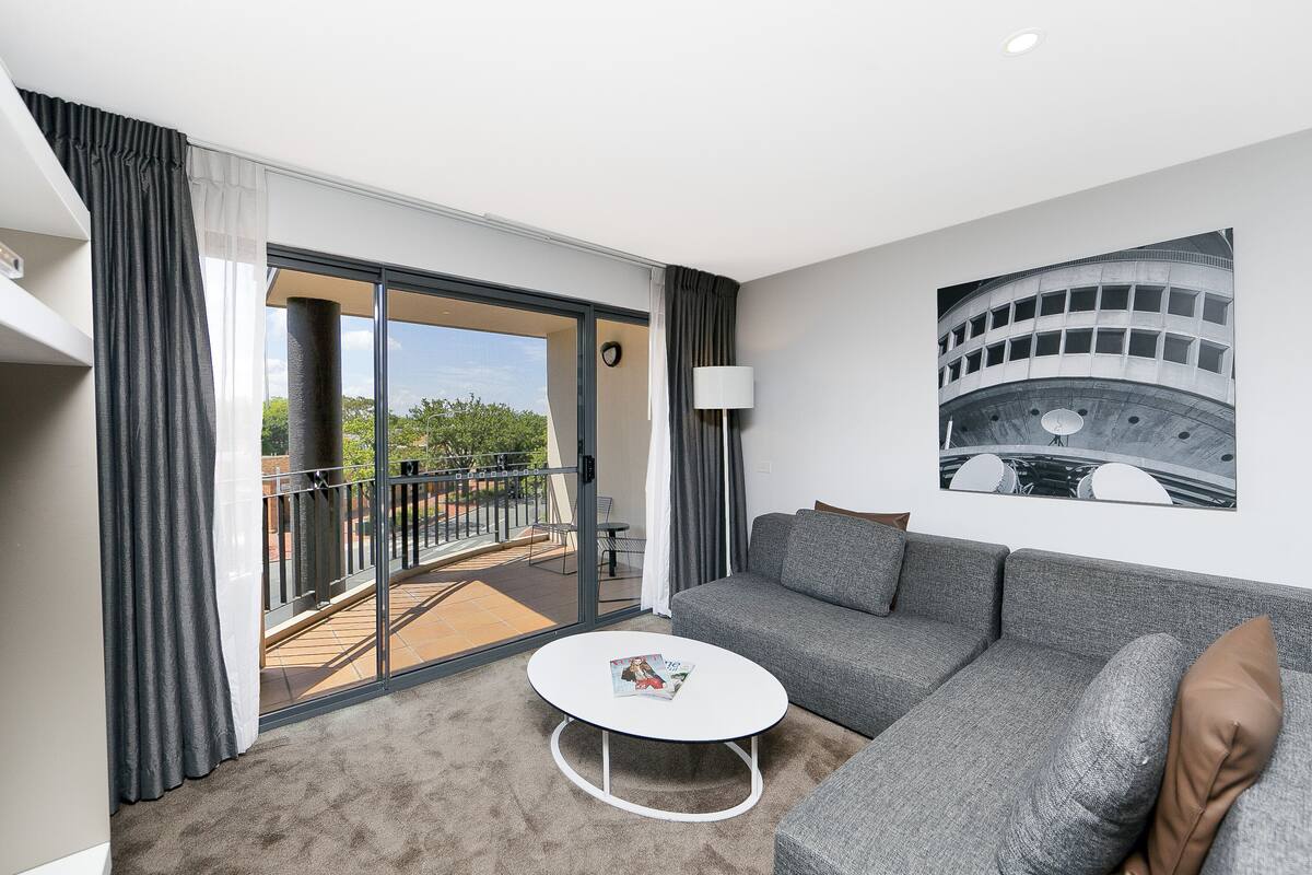 Un salon moderne avec un canapé gris, une table basse blanche et un balcon offrant une vue sur la verdure.