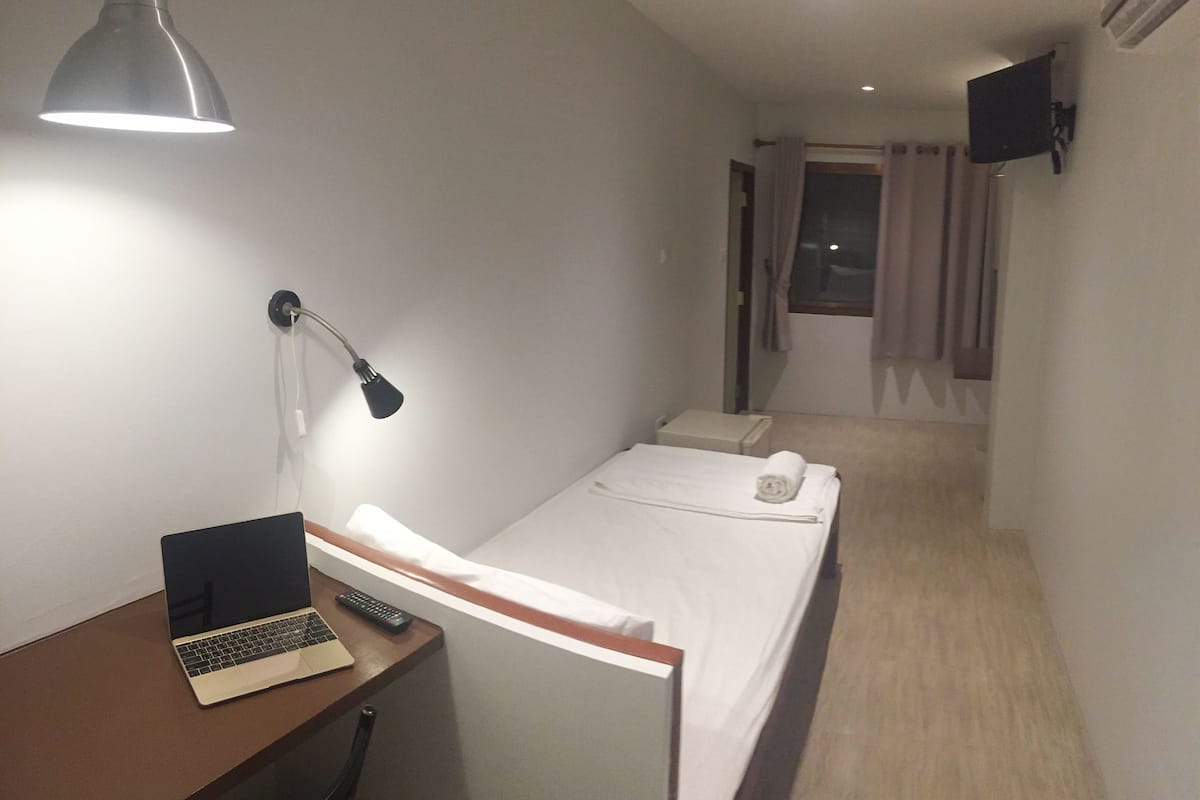 A small hotel room with a single bed, a desk with a laptop, a desk lamp, and a TV mounted on the wall.