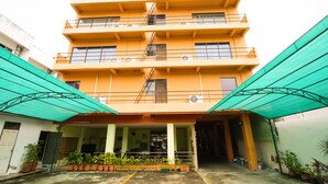 Un edificio de varios pisos con techo plano, balcones y una zona de entrada cubierta con plantas en macetas.