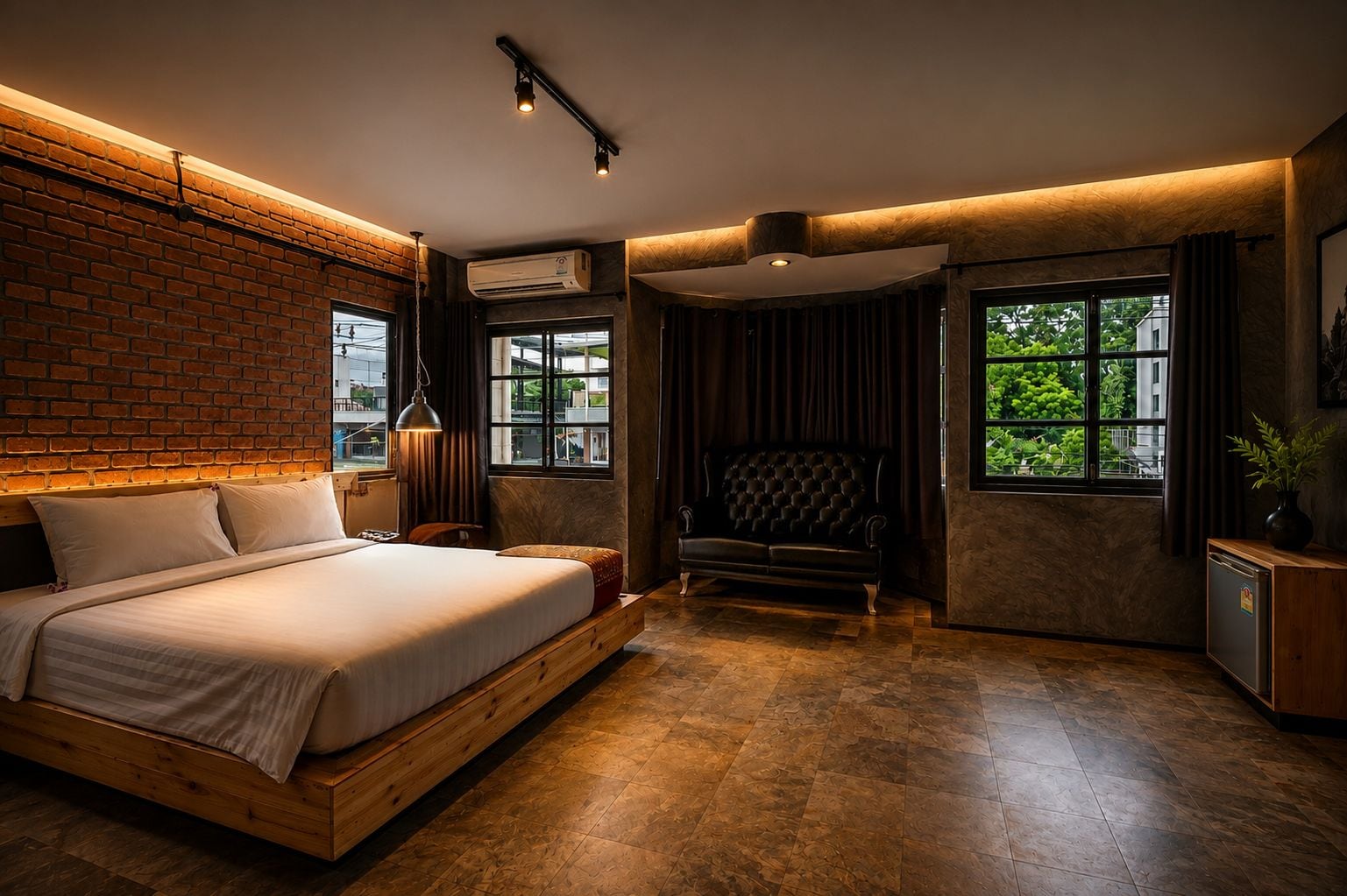 A modern bedroom with a brick wall, a large bed, a leather sofa, and a view of greenery through the windows.