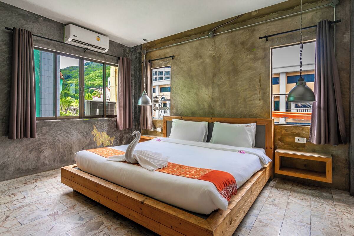 A bedroom with a wooden bed, a nightstand, a hanging lamp, and a view of greenery outside.