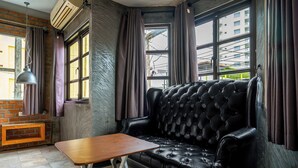 Un sofá de cuero negro, una mesa de madera y una lámpara colgante en una habitación con ventanas amplias y una pared de ladrillo.