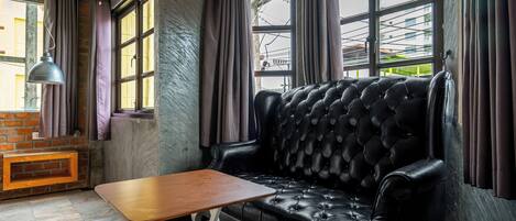 A black leather sofa, a wooden table, and a hanging lamp in a room with large windows and a brick wall.