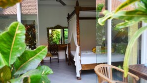 A room with a four-poster bed, a wooden chair, a table, and a view of greenery through a glass door. - Aquarius Beach Hotel (Denpasar)
