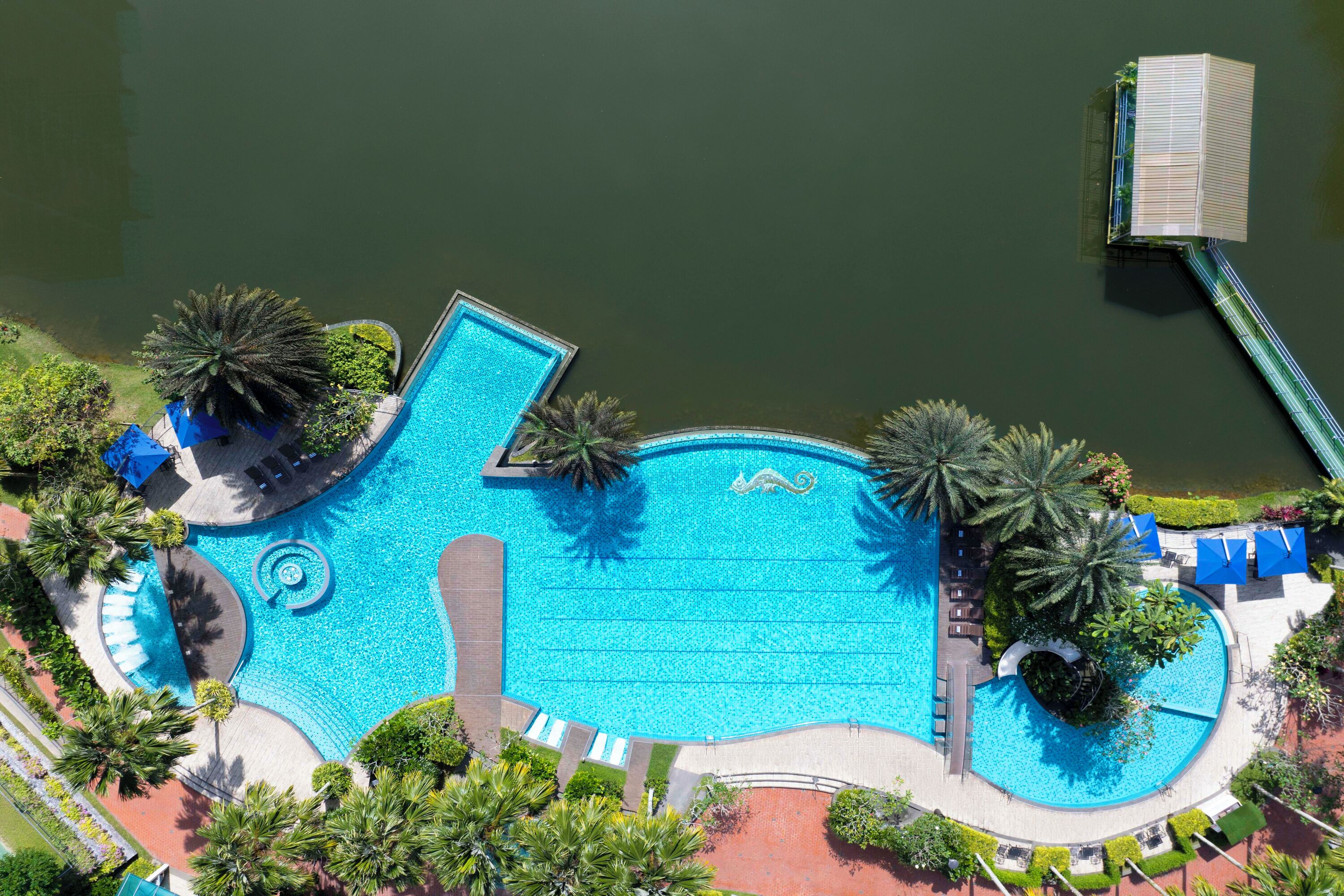An aerial view of a swimming pool with a bridge, palm trees, and lounge areas.
