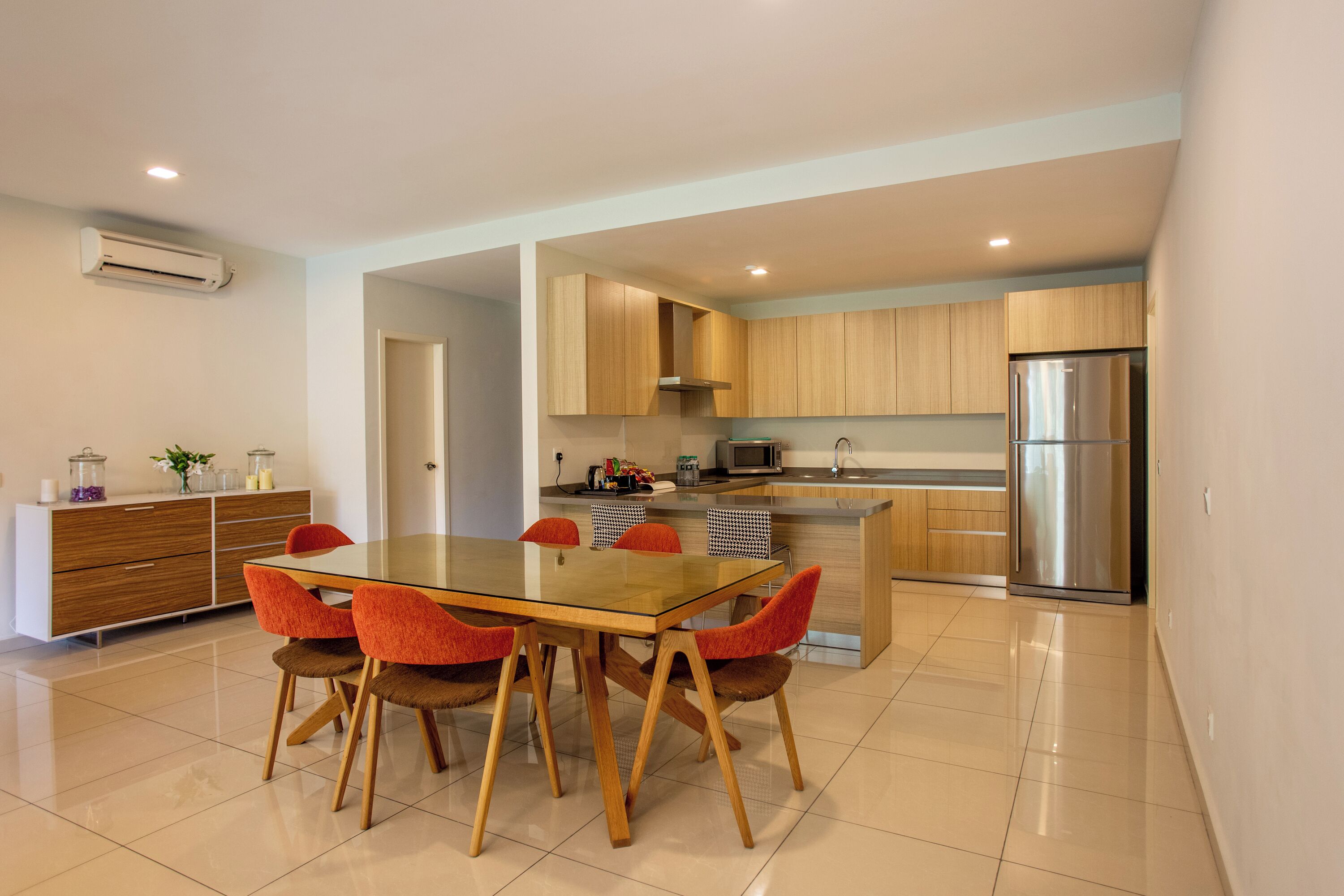 A modern kitchen with wooden cabinets, a stainless steel refrigerator, and a glass dining table with orange chairs.
