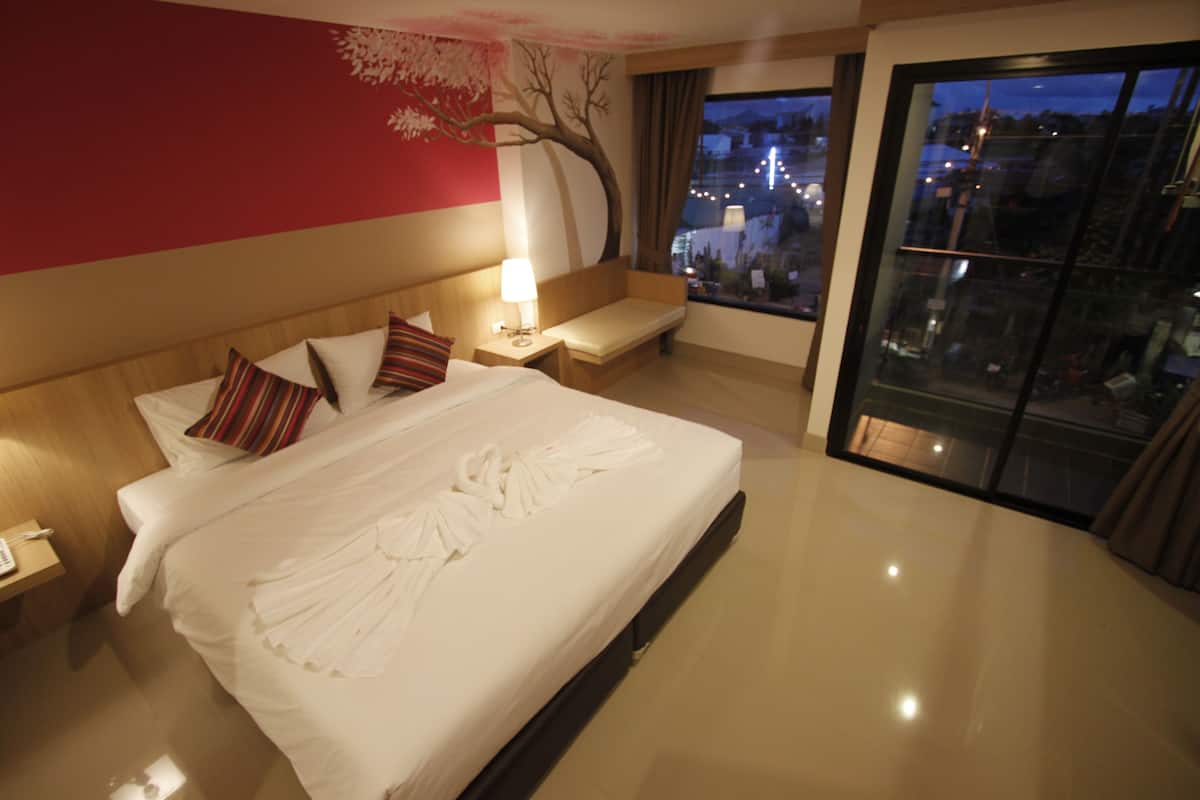 Ein Hotelzimmer mit einem großen Bett, einem Nachttisch, einer Lampe, einer Bank und Blick auf die Stadt durch ein großes Fenster.