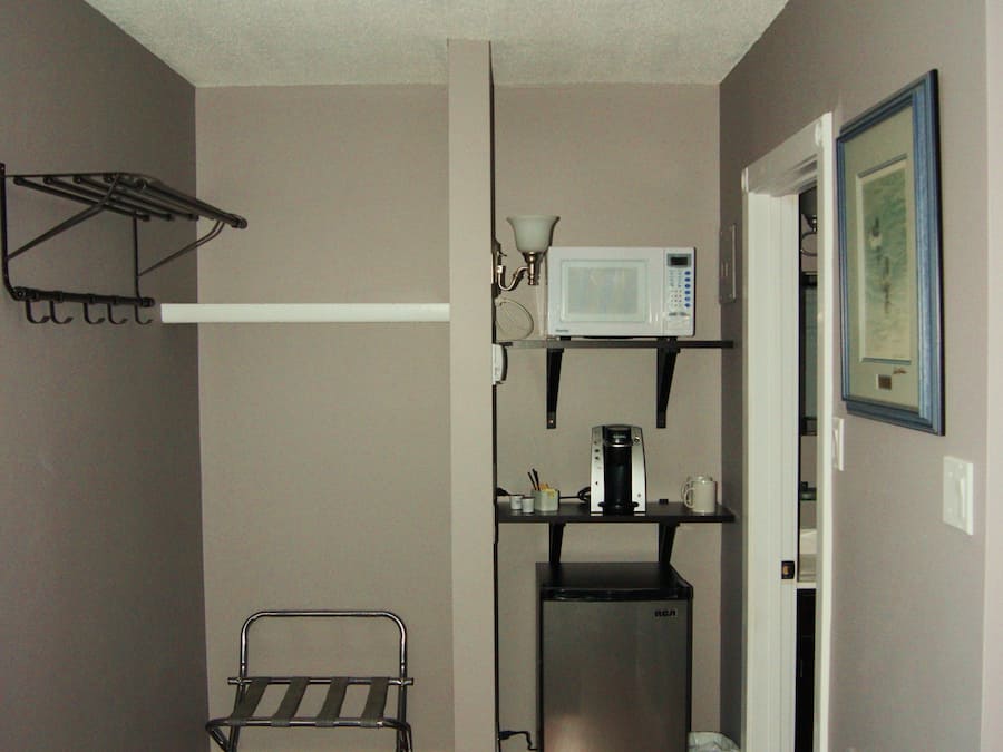 Cocina compacta con microondas, cafetera y un refrigerador pequeño. Un estante metálico está sujeto a la pared, y se observa un cesto de basura.