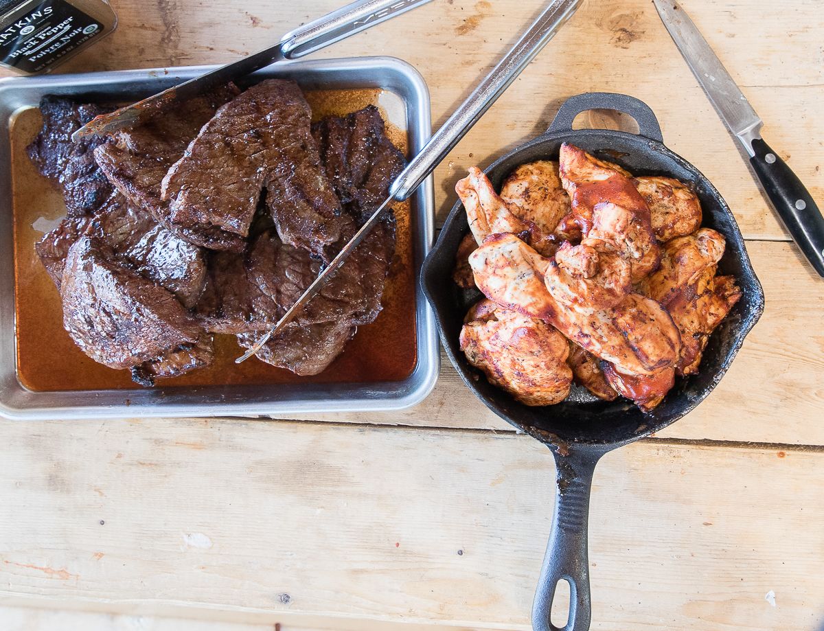 A tray of cooked meat and a skillet with chicken pieces on a wooden surface.
