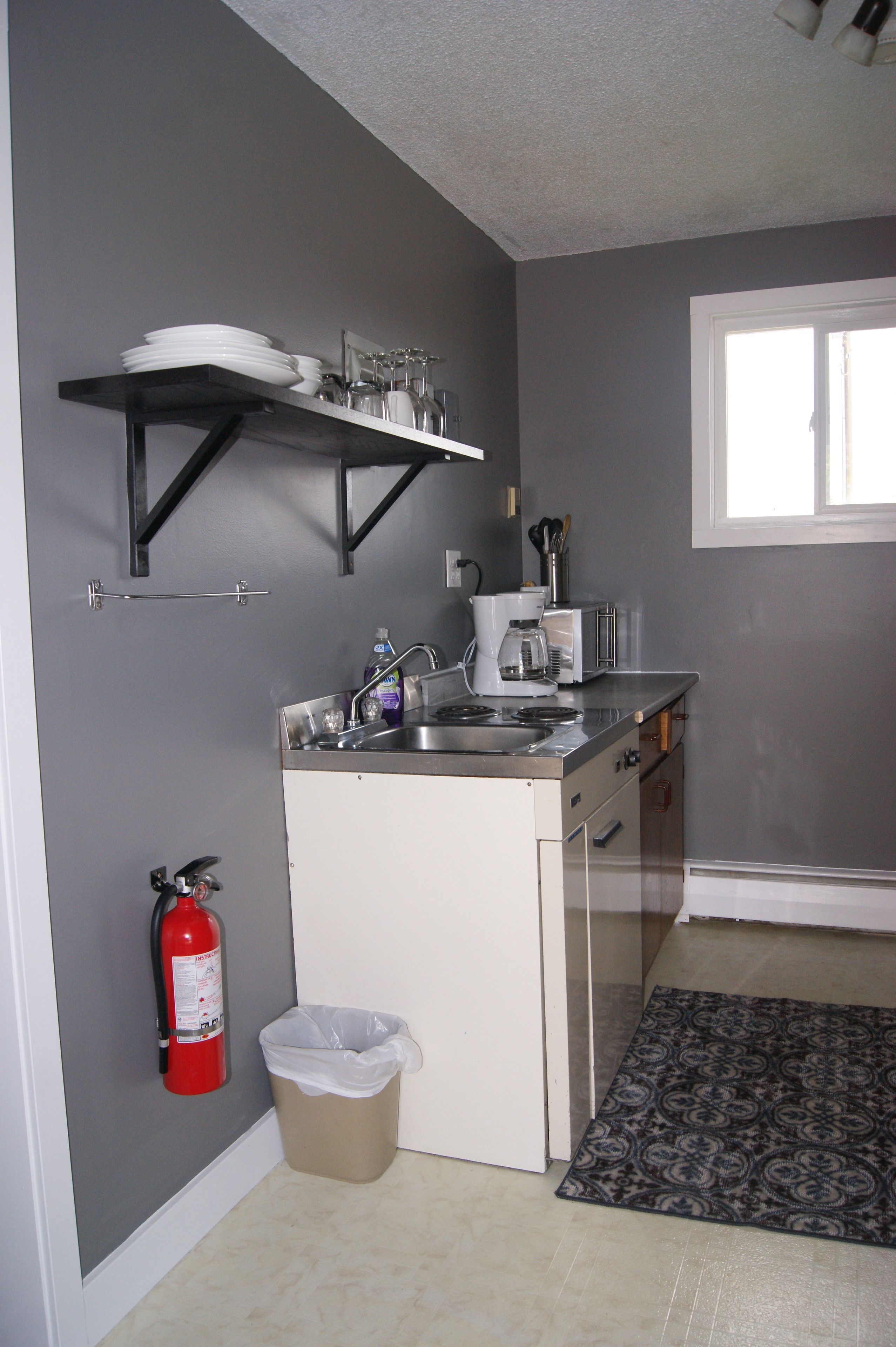 A compact kitchen with a built-in stove, a sink, and a small countertop. A fire extinguisher is mounted on the wall, and a trash bin is placed near the sink.