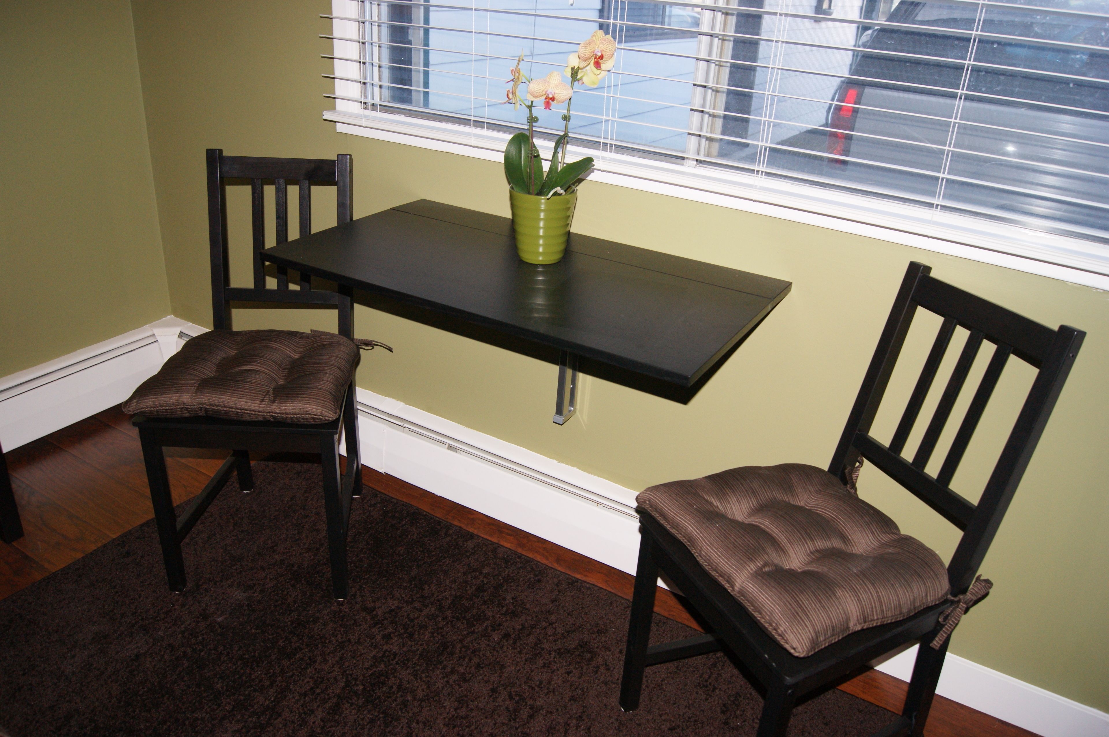 A dining area with a table and two chairs, a green vase with flowers, and a window with blinds.