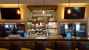 Un área de bar con un letrero que dice "GENE'S QUARTERS" sobre una estantería llena de botellas de licor. Hay cuatro taburetes de bar frente al mostrador.