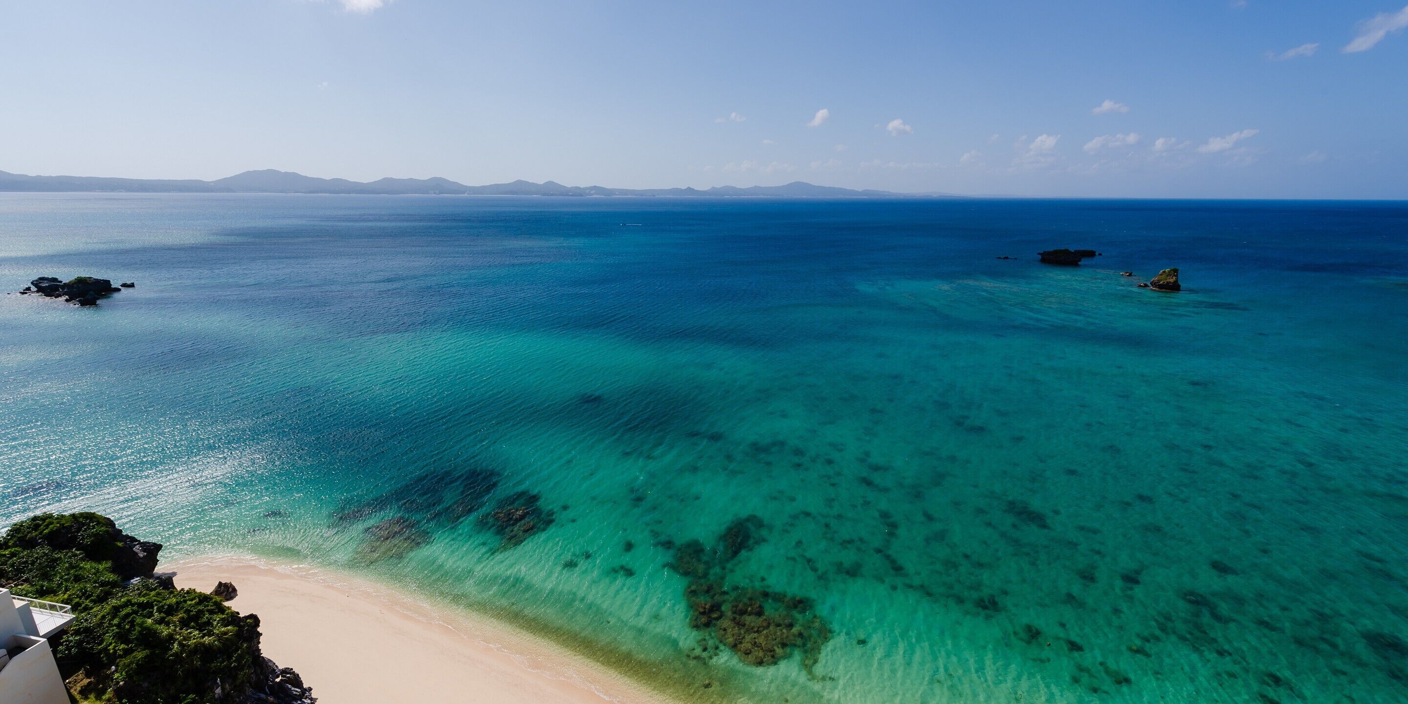 Une vue sur la côte, avec une eau turquoise et limpide, une plage de sable et des reliefs terrestres qui se dessinent à l’horizon.