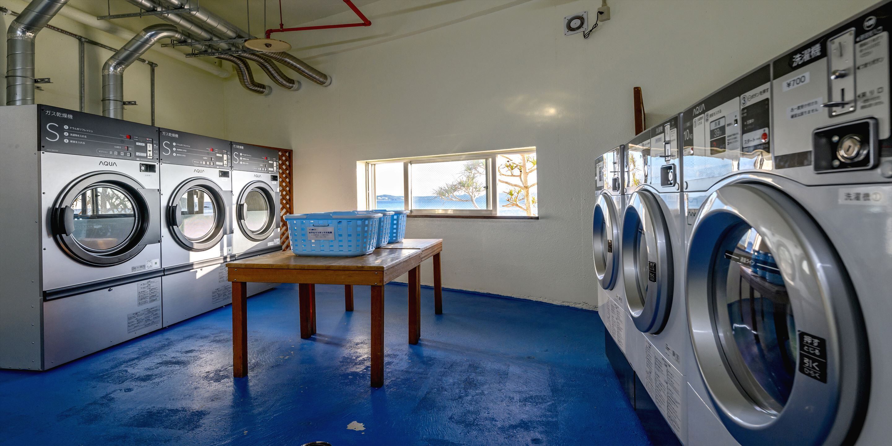 A laundry room with two rows of washing machines, a table with baskets, and a window with a view.