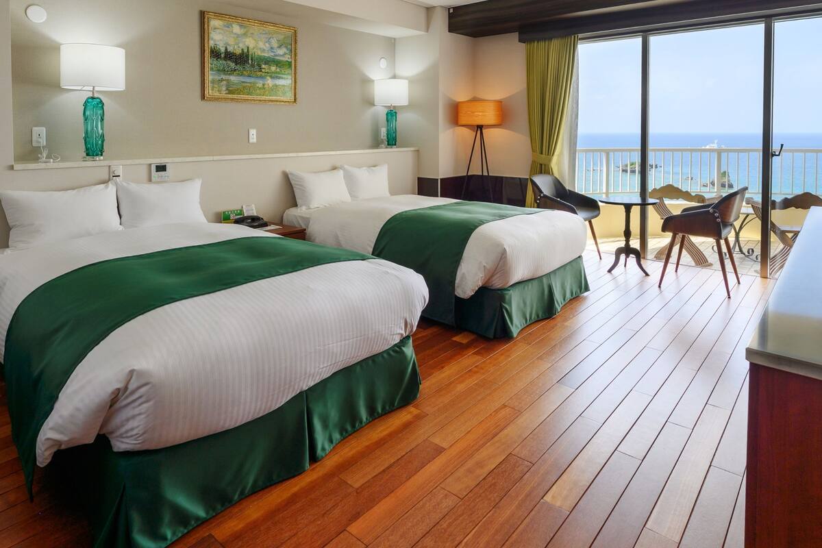 A hotel room with two beds, a balcony with a view of the sea, and a desk with a computer.