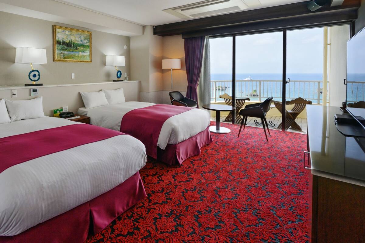 A hotel room with two beds, a red carpet, a balcony with a view of the sea, and a desk with a chair.