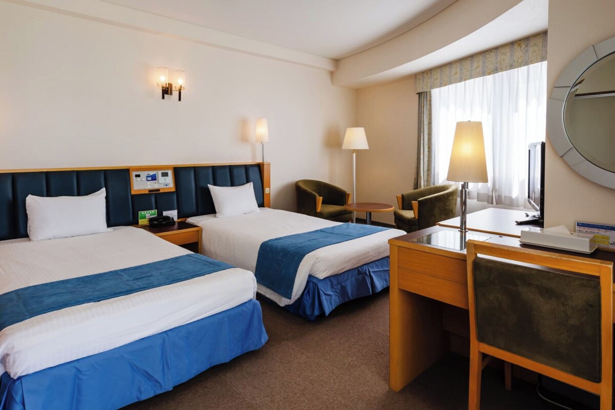 A hotel room with two beds, a desk, a chair, a lamp, a bedside table, and a wall-mounted TV.