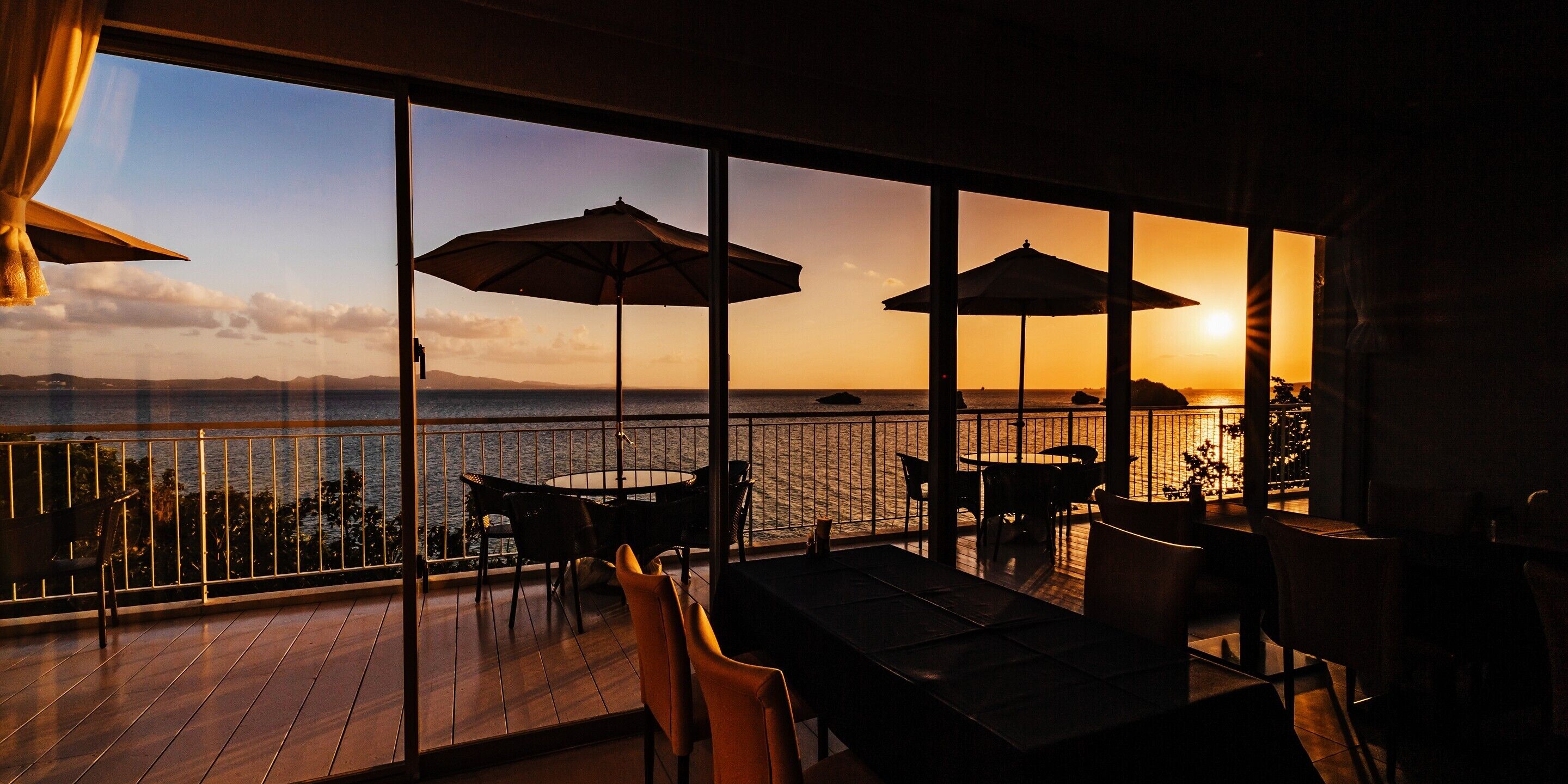 A terrace with tables and chairs, large umbrellas, and a view of the sunset over the water.