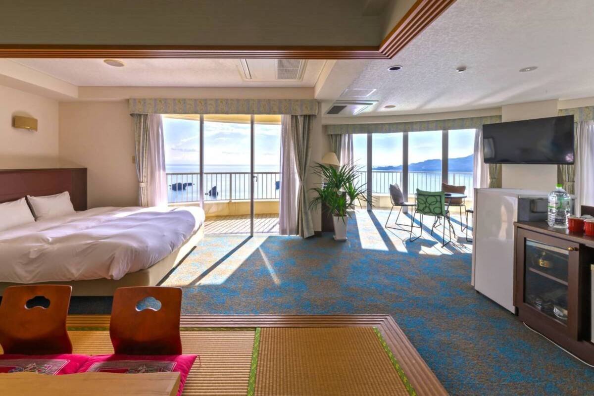 A hotel room with a bed, a seating area with a table and chairs, a television, and a balcony with a view of the sea.