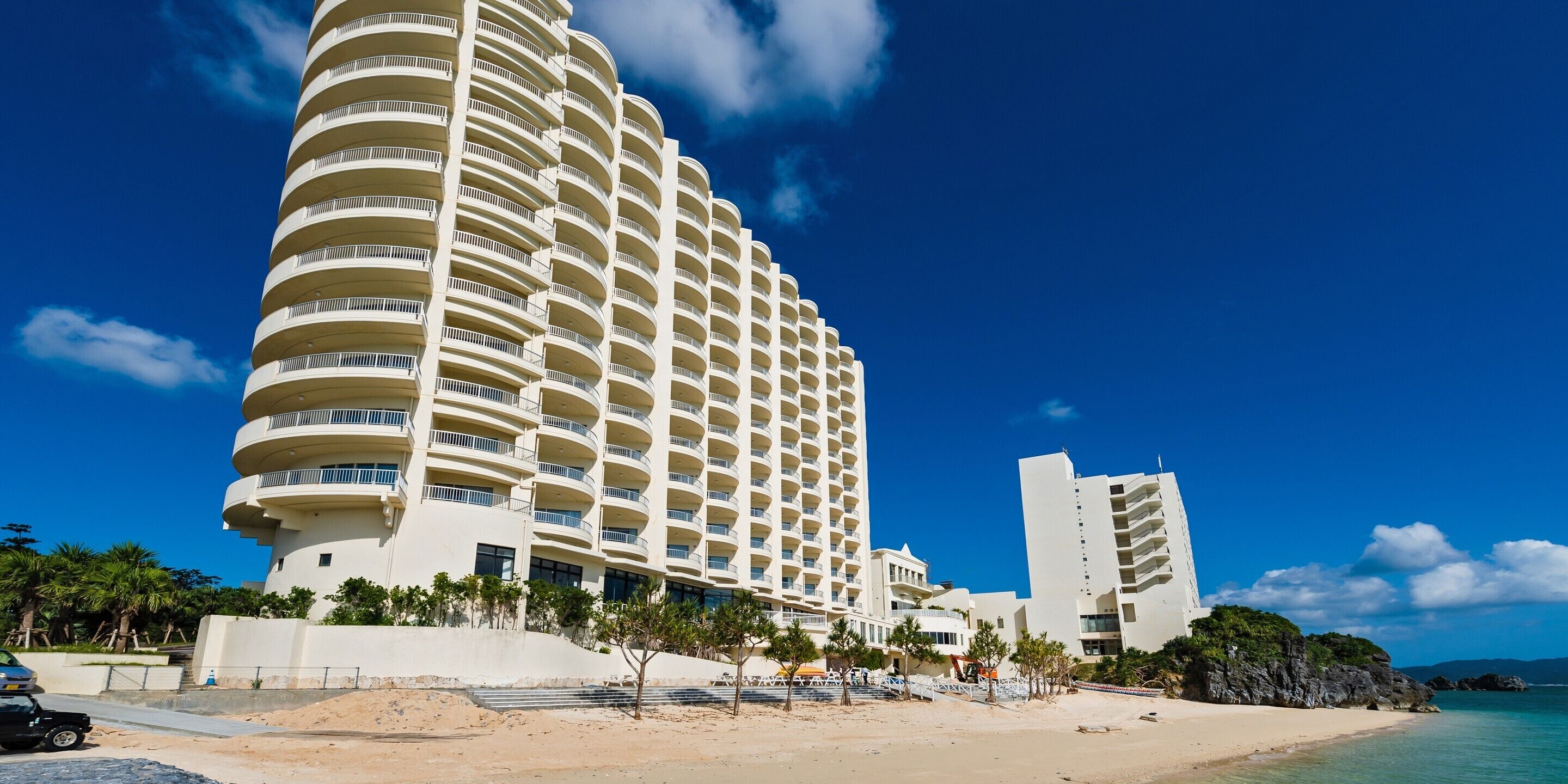 A multi-story hotel with balconies, located near a beach with clear skies.
