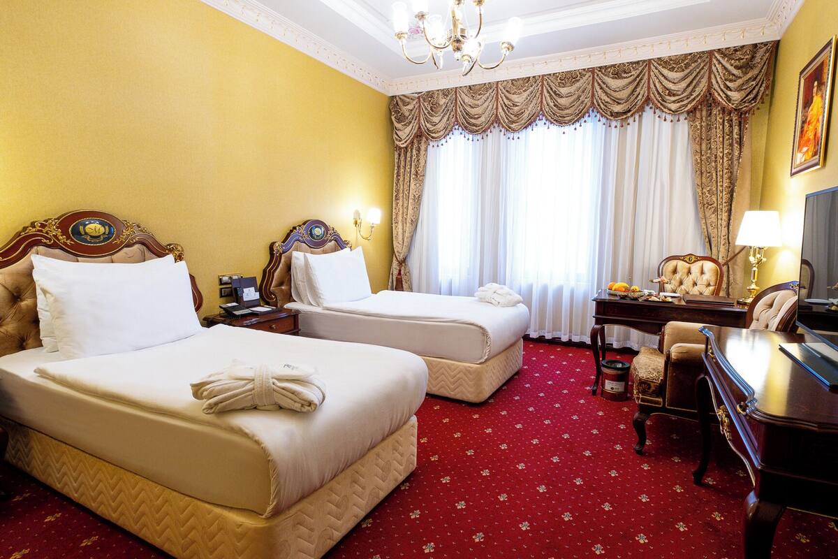 A hotel room with two beds, a desk with a chair, a television, and a large window with curtains.