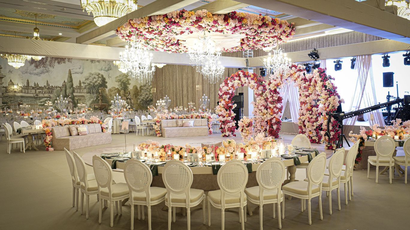 A banquet hall with round tables, white chairs, floral arrangements, chandeliers, and a stage.
