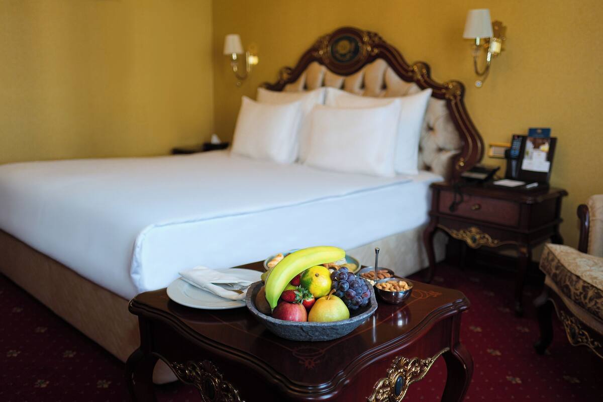 A neatly made bed with white linens, a fruit bowl on a side table, a chair, and a nightstand with a lamp.