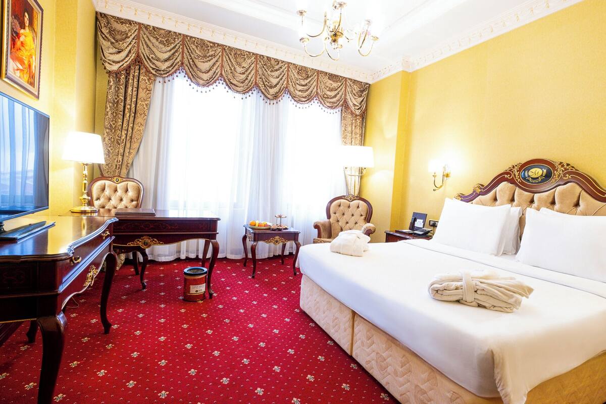 A hotel room with a large bed, a desk, two chairs, a television, and a chandelier.
