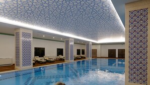 A large indoor swimming pool with a geometric ceiling design, lounge chairs, and a tiled wall with a detailed pattern. - Meyra Palace (Ankara)