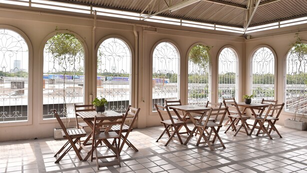 A covered outdoor seating area with multiple tables and chairs, large arched windows, and a view of the cityscape. - G9 Bangkok (Bangkok)