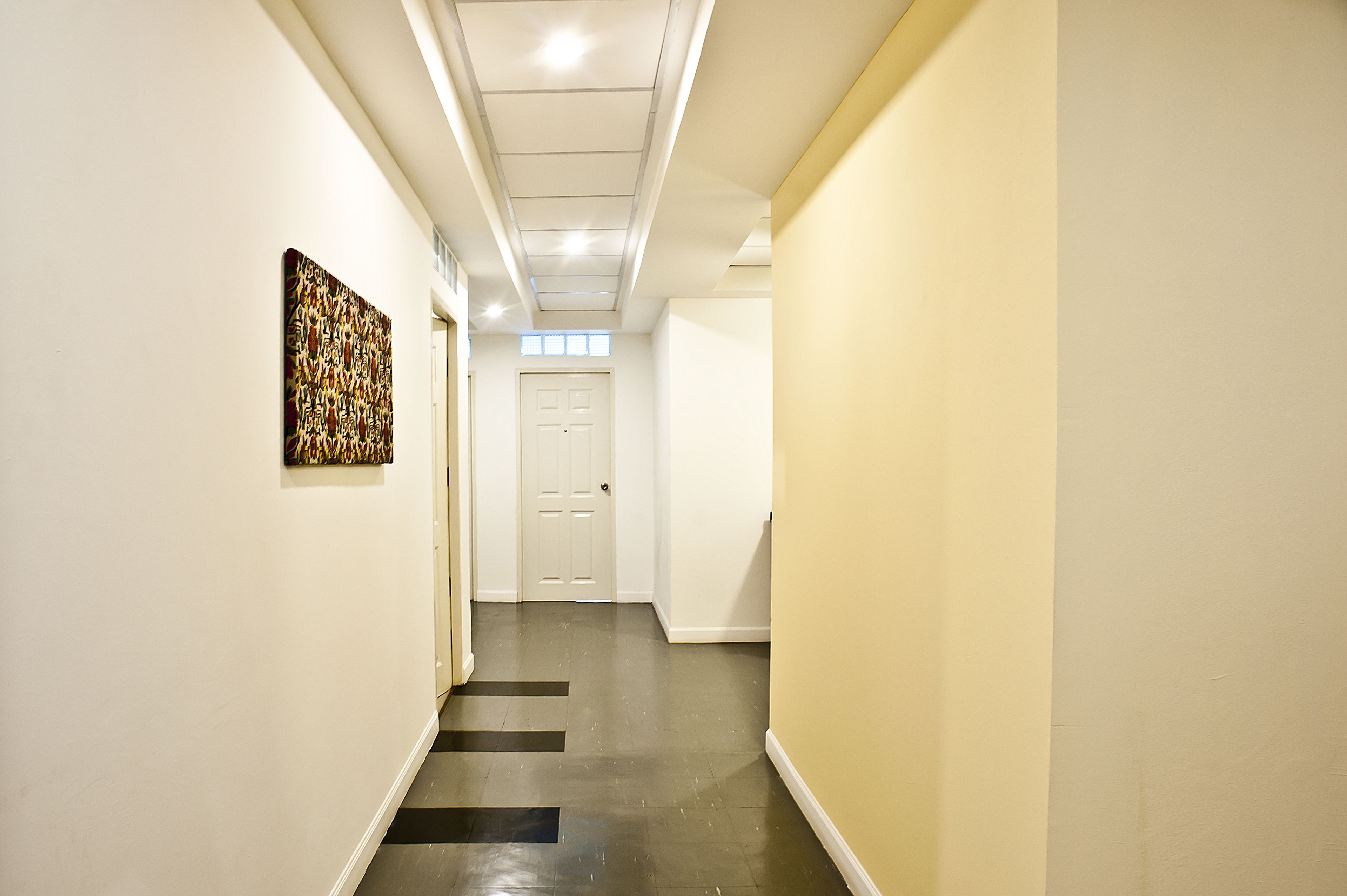 A hallway with a white door at the end, a black and white floor, and a painting on the left wall.