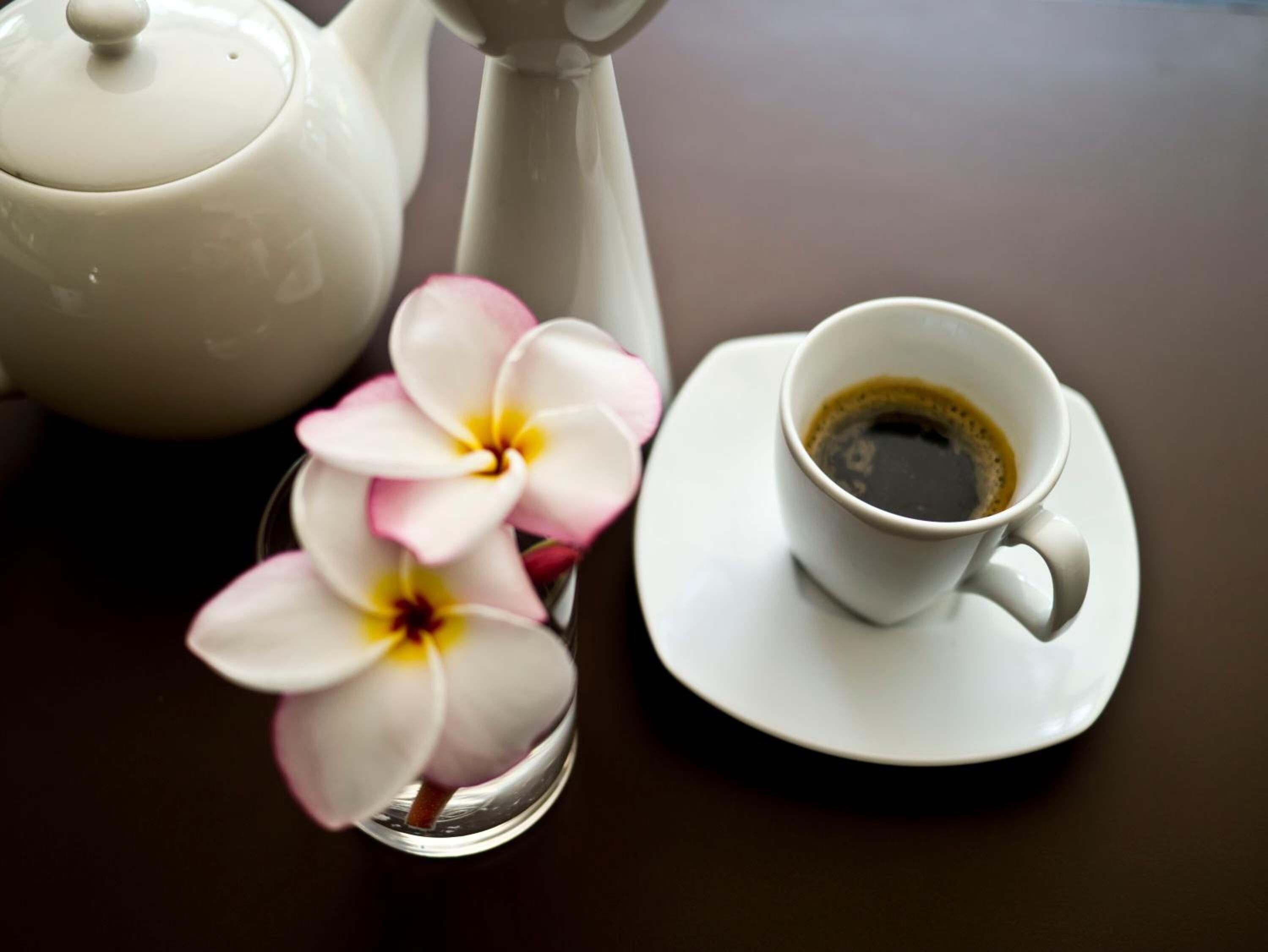 A white teapot, a cup of black coffee, and a spoon with white and pink flowers on a dark table.