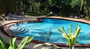 A swimming pool with a curved design, surrounded by lush greenery and a few lounge chairs. - Granpa's Inn Hotel Bougainvillea (Anjuna)