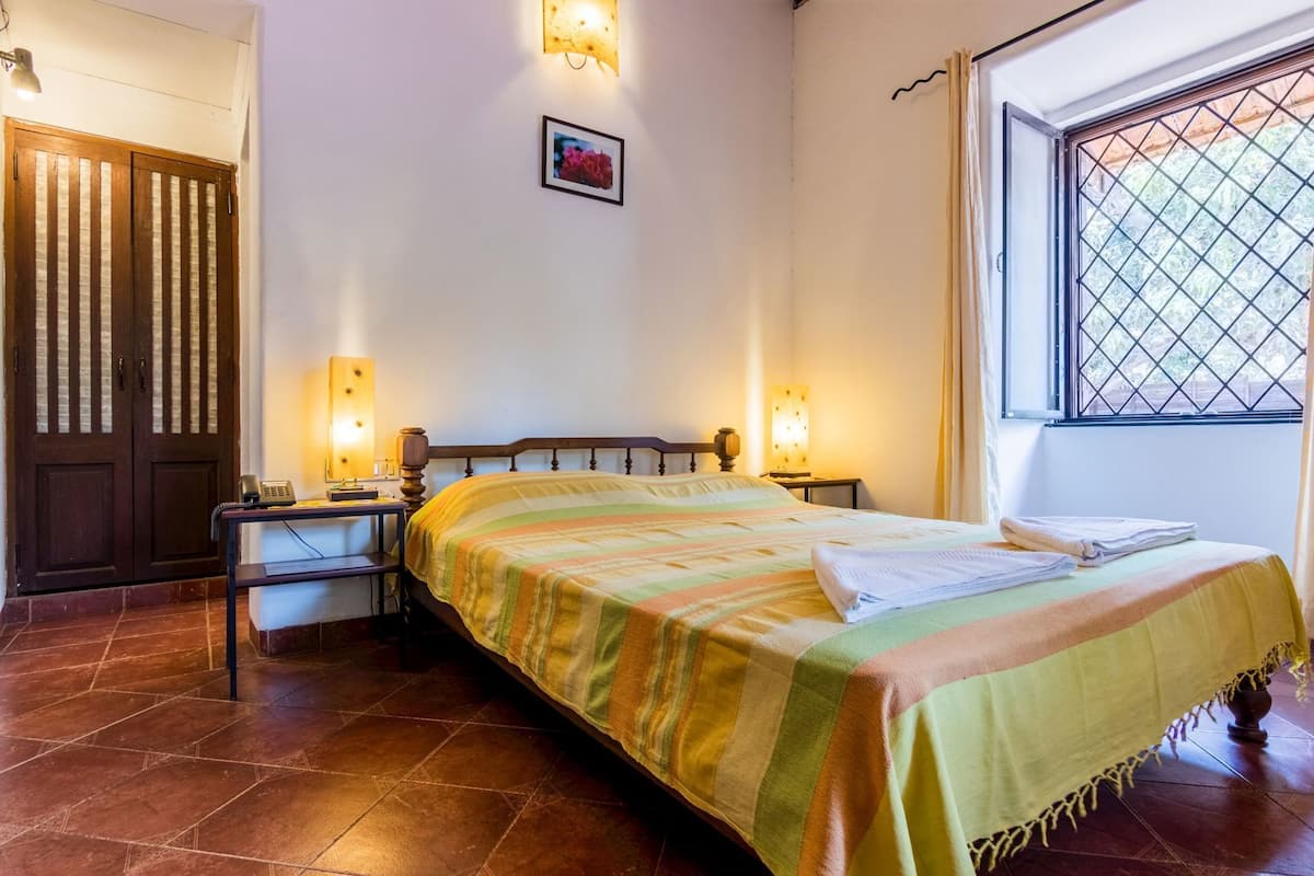 A room with a bed, two bedside tables, a desk, a chair, a door, a window with a view, and a wall-mounted picture.