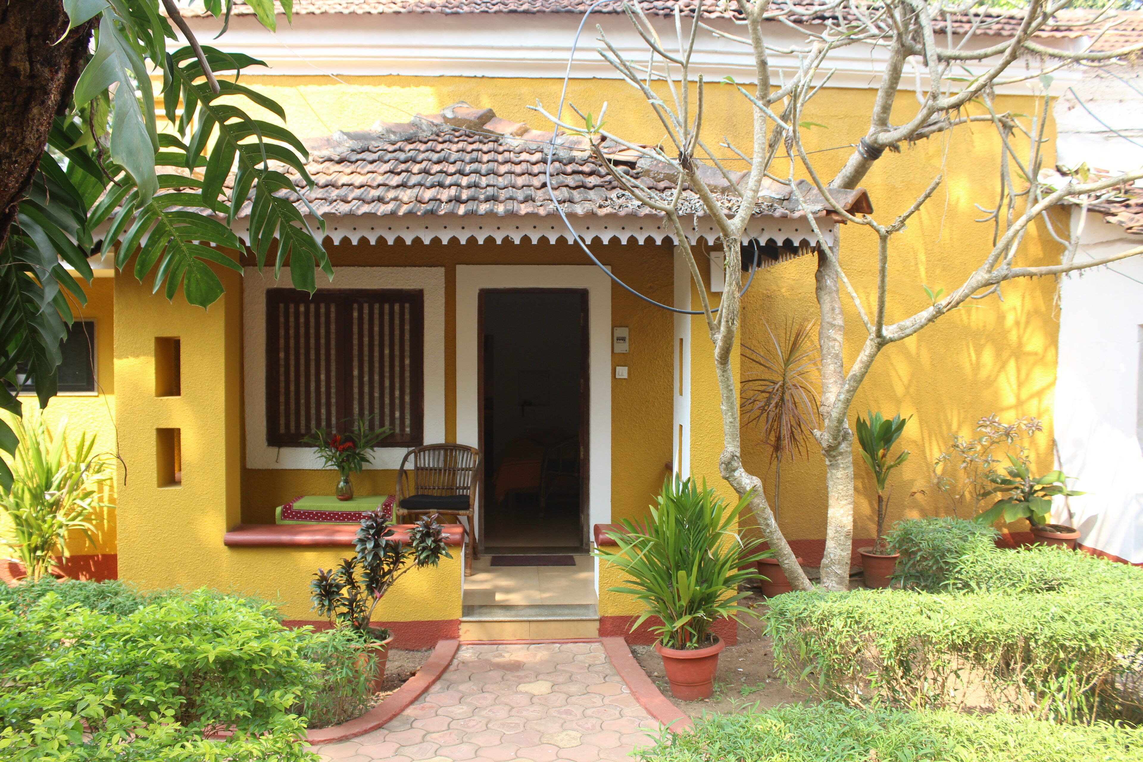 A yellow building with a tiled roof, a wooden door, and a small porch with a chair and a table. There are potted plants around the building.