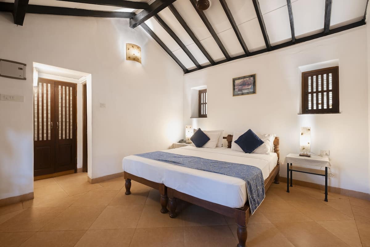 A bedroom with a large bed, a nightstand, a wall-mounted lamp, and a door with vertical blinds.