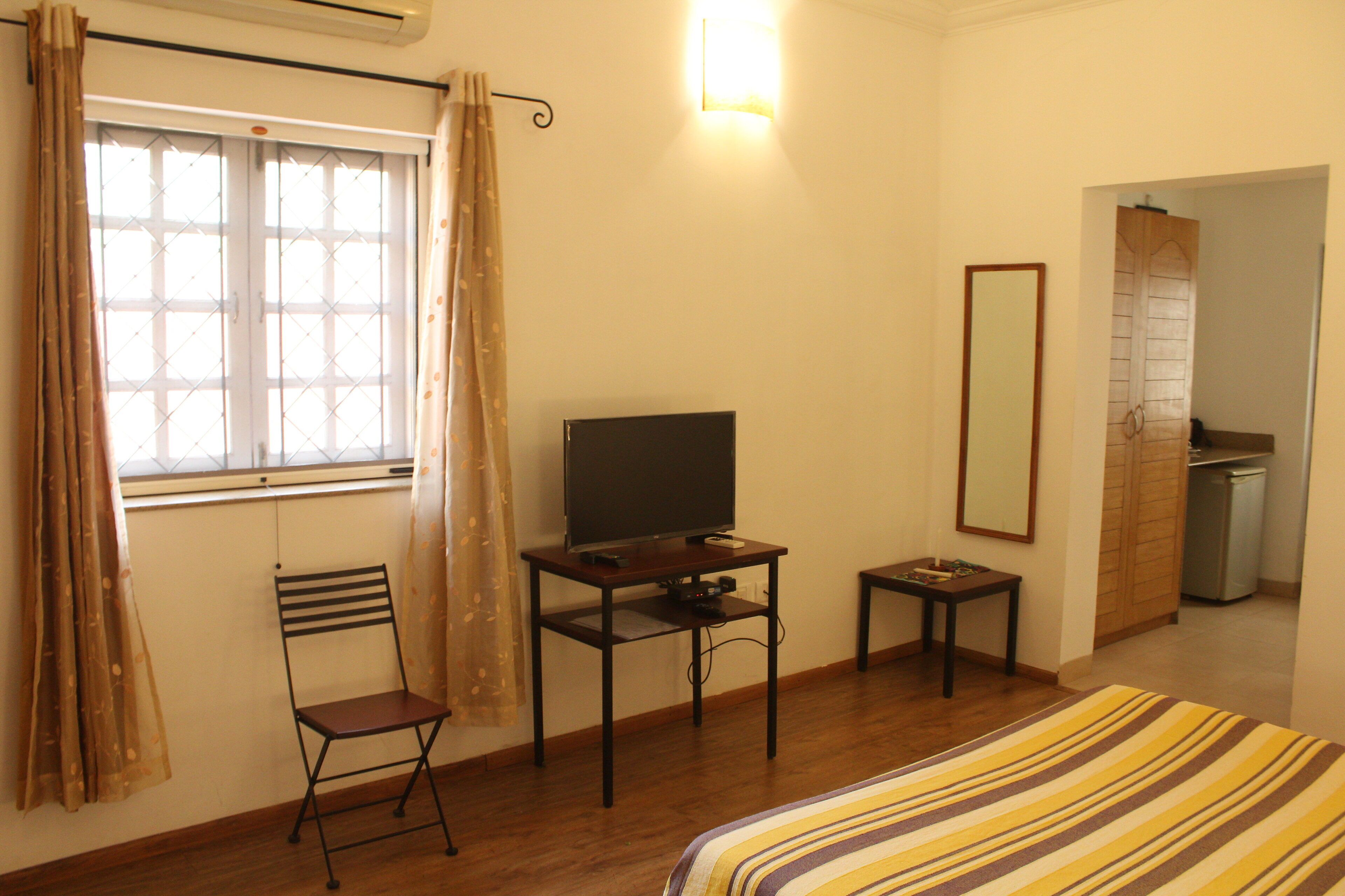 Una habitación con cama, televisión sobre un mueble bajo, una silla, una mesita y un espejo en la pared.