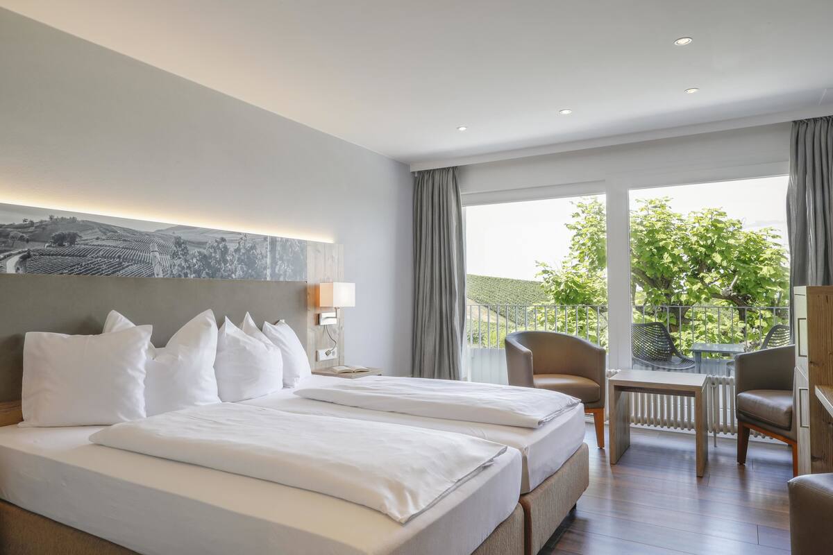 A hotel room with two beds, a desk, chairs, and a view of greenery outside.