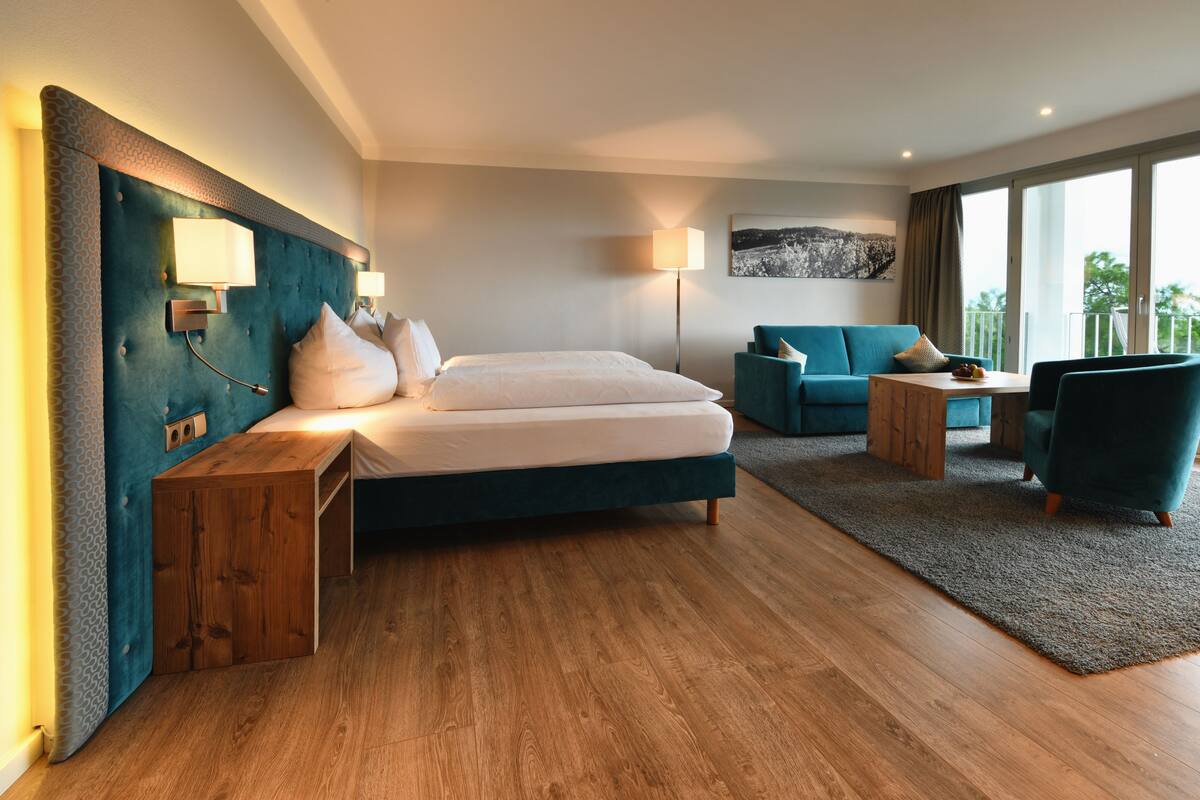A modern hotel room with a large bed, a seating area with a sofa and armchairs, a wooden side table, and a large window offering a view.