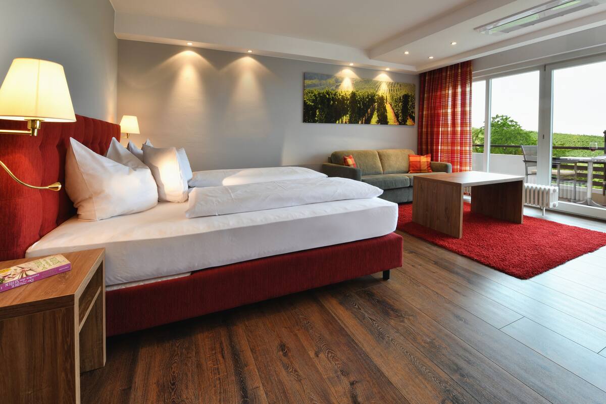 A modern hotel room with a large bed, a sofa, a small table, and a view of the outdoors.