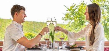 Um casal brindando com taças de vinho à mesa, com vista para um vinhedo.