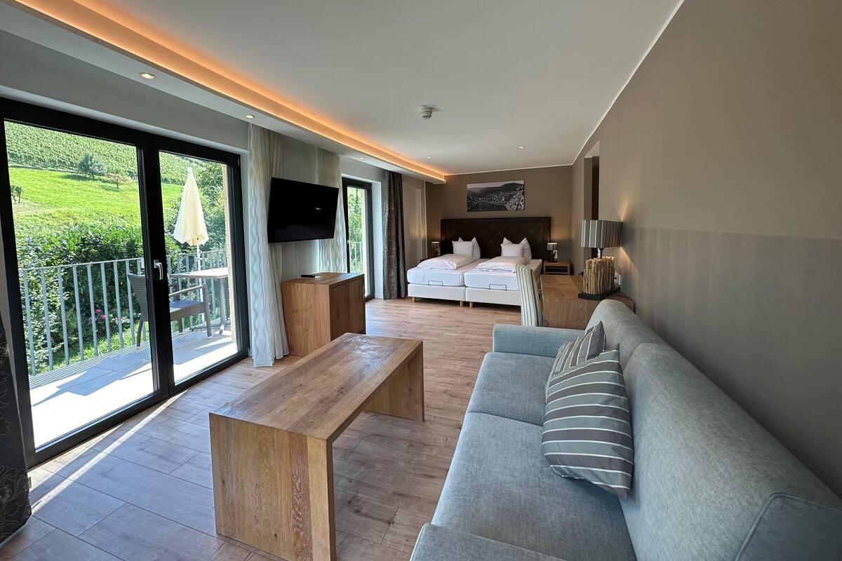 A modern hotel room with a large bed, a sofa, a TV, and a balcony with a view of greenery.