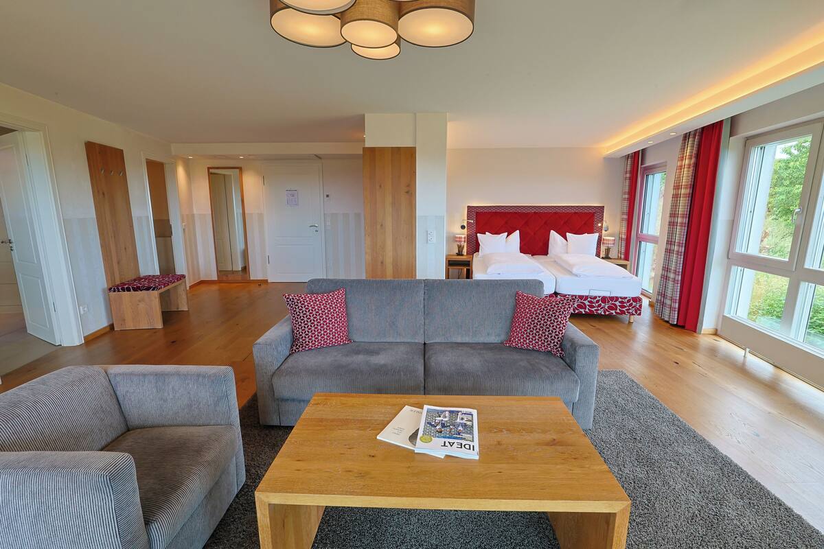 A hotel room with a large bed, a grey sofa, a wooden coffee table, and a view of greenery through the windows.
