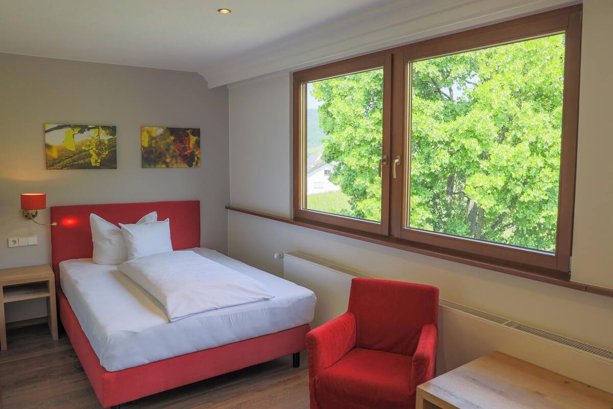 A bedroom with a bed, a red chair, a small table, and a window with a view of trees.