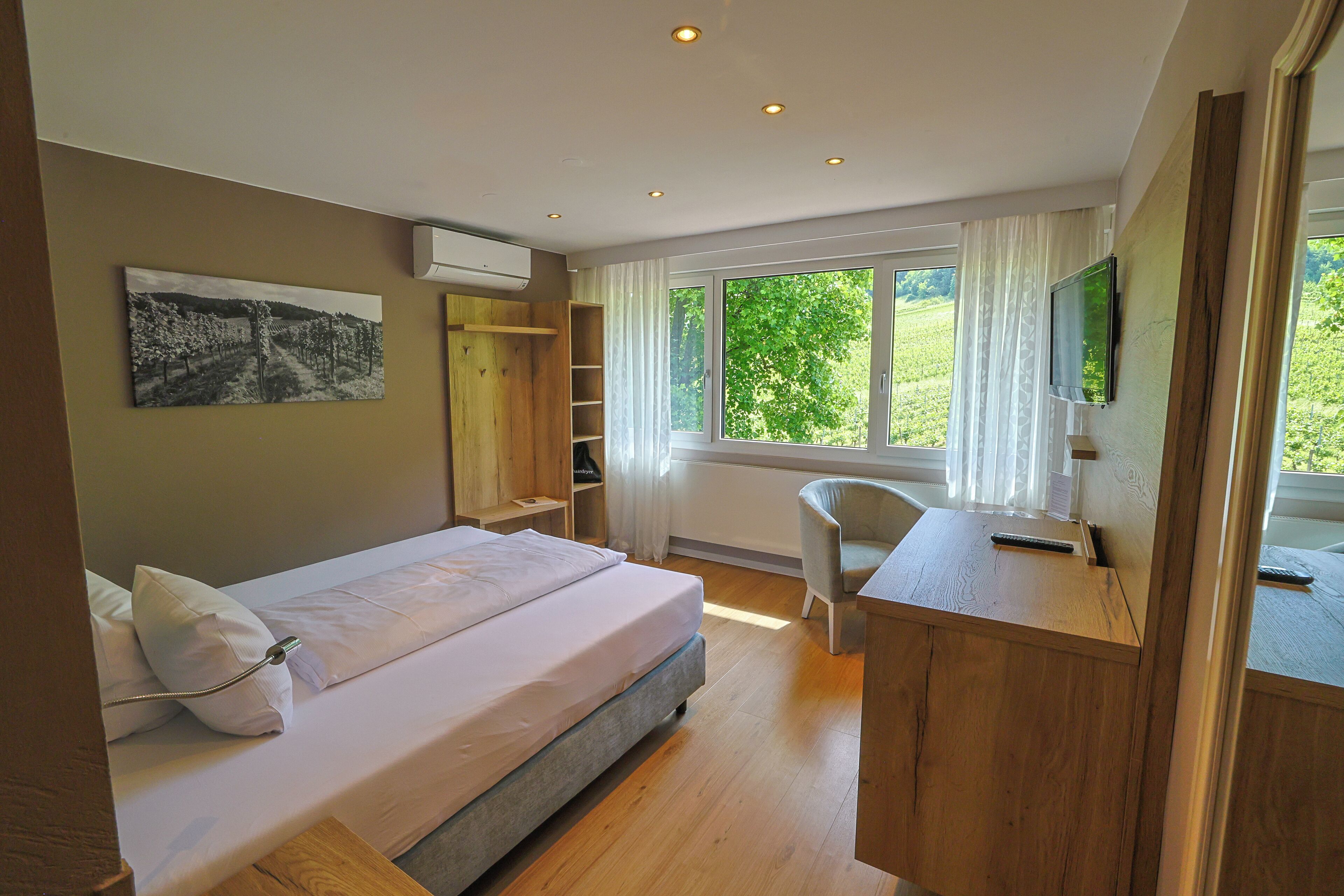 A bedroom with a bed, a desk, a chair, a TV, and a window with a view of greenery.